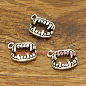 May include: Three silver-colored, open-mouthed charm pendants shaped like vampire teeth. Each charm has a small loop at the top for attaching to jewelry. The charms are displayed on a wooden surface.