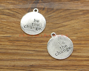 May include: Two silver charm pendants with the words "be the change." on each. The charms are round and have a hole at the top for attaching to a chain or bracelet.
