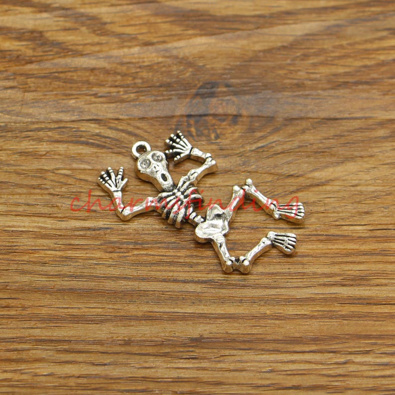 May include: Silver-toned skeleton charm with a detailed design. The skeleton is in a pose of surprise or fear, with its arms raised and its mouth open.