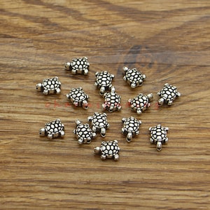 Op de afbeelding: Een verzameling van 15 zilveren schildpadbedels, elk met een gedetailleerde, getextureerde schelp en vier kleine pootjes. De bedels zijn verspreid over een houten oppervlak.