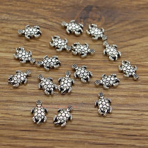 Puede incluir: Pequeños abalorios plateados con forma de tortuga y patrones detallados en el caparazón. Estos abalorios están dispersos sobre una superficie de madera, mostrando su intrincado diseño, ideal para bisutería.