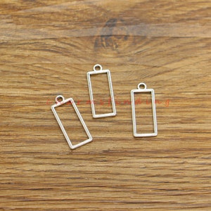 Op de afbeelding: Drie rechthoekige zilveren metalen hangerframes met een lus aan de bovenkant. De frames liggen op een houten oppervlak.