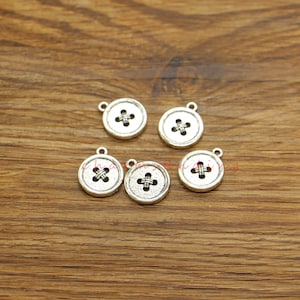 May include: Five silver-toned metal button charms with a cross-stitch design. The charms are round and have a hole at the top for attaching to jewelry or other crafts.