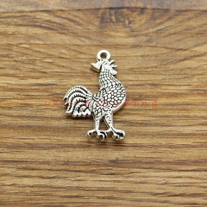 Può includere: Ciondolo in argento a forma di gallo. Il gallo è in piedi con le ali spiegate e la testa alta. Il ciondolo è dettagliato con una superficie testurizzata.