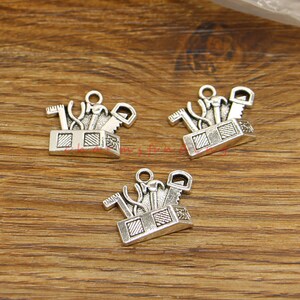May include: Three silver-toned metal charms shaped like toolboxes with various tools inside. Each charm has a loop at the top for attaching to a necklace or bracelet.