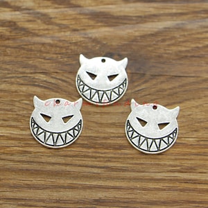 May include: Three silver-toned metal charms with a devil face design. Each charm features a smiling face with sharp teeth, triangular eyes, and small horns. The charms are round with a small hole at the top for stringing.