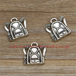 May include: Three silver charm pendants shaped like backpacks with straps and buckles. The charms are detailed with a pocket on the front and are suitable for jewelry making.