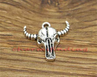 May include: A silver-toned metal charm shaped like a bull skull with prominent horns and a small circular loop at the top for attachment. The surface features dark recessed detailing to highlight the anatomical structure of the animal skull.