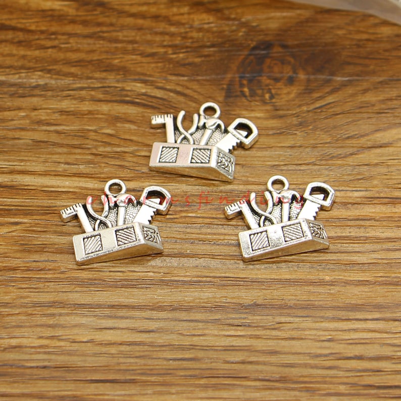 May include: Three silver charm pendants shaped like toolboxes with various tools inside. The toolboxes are detailed with a textured surface and a small loop at the top for attaching to a chain or bracelet.