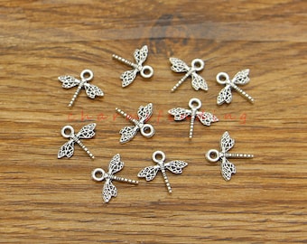 Könnte beinhalten: Silberfarbene Libellen-Anhänger mit einer Öse zum Anbringen an Schmuck oder Bastelarbeiten. Die Anhänger sind etwa 2,5 cm lang und haben ein detailliertes Design.