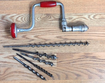 May include: An antique hand drill with a red handle and a set of drill bits. The drill is made of metal and has a silver finish. The drill bits are various sizes and shapes, and are made of metal. The items are on a wooden surface.