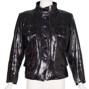 Op de afbeelding: Zwarte jas met slangenleertextuur, opstaande kraag, vier voorzakken en knoopsluitingen. De jas wordt getoond op een mannequin tegen een witte achtergrond. De glanzende afwerking van de jas draagt bij aan de luxueuze uitstraling.