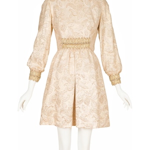 May include: A short, cream-colored dress with a gold floral pattern. The dress has a gold beaded belt and gold trim on the cuffs.