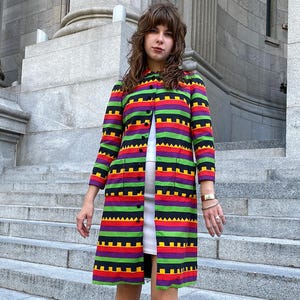 May include: A knee-length coat featuring a vibrant horizontal striped pattern in bright green, red, purple, and black. The coat has a high neckline, long sleeves, a front button closure, and two rectangular patch pockets on the front panels.