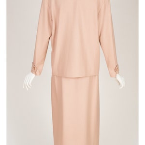 May include: A peach-coloured skirt suit on a mannequin. The suit features a long-sleeved jacket with shoulder pads and a knee-length skirt. The jacket has buttoned cuffs and a collared neckline. The mannequin is on a clear glass stand.