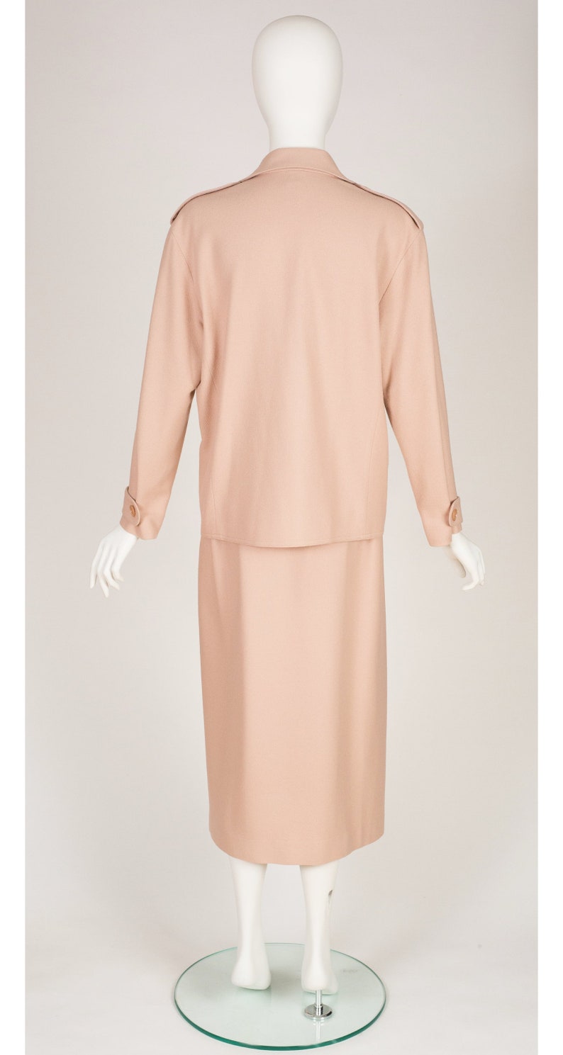May include: A peach-coloured skirt suit on a mannequin. The suit features a long-sleeved jacket with shoulder pads and a knee-length skirt. The jacket has buttoned cuffs and a collared neckline. The mannequin is on a clear glass stand.