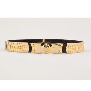 May include: Gold-tone metal belt with a black elastic band. The belt features a central gold-tone plate with a circular design and is secured with a clasp. The belt's design includes a series of gold-tone metal segments.