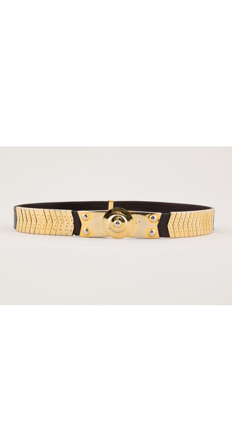 May include: Gold-tone metal belt with a black elastic band. The belt features a central gold-tone plate with a circular design and is secured with a clasp. The belt's design includes a series of gold-tone metal segments.