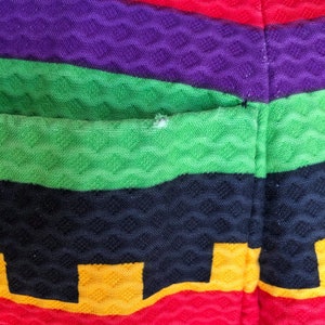 May include: A textured fabric garment featuring horizontal stripes in vibrant shades of yellow, red, purple, green, and black. The material has a raised, diamond-shaped waffle pattern throughout, with a small visible snag on the green stripe.