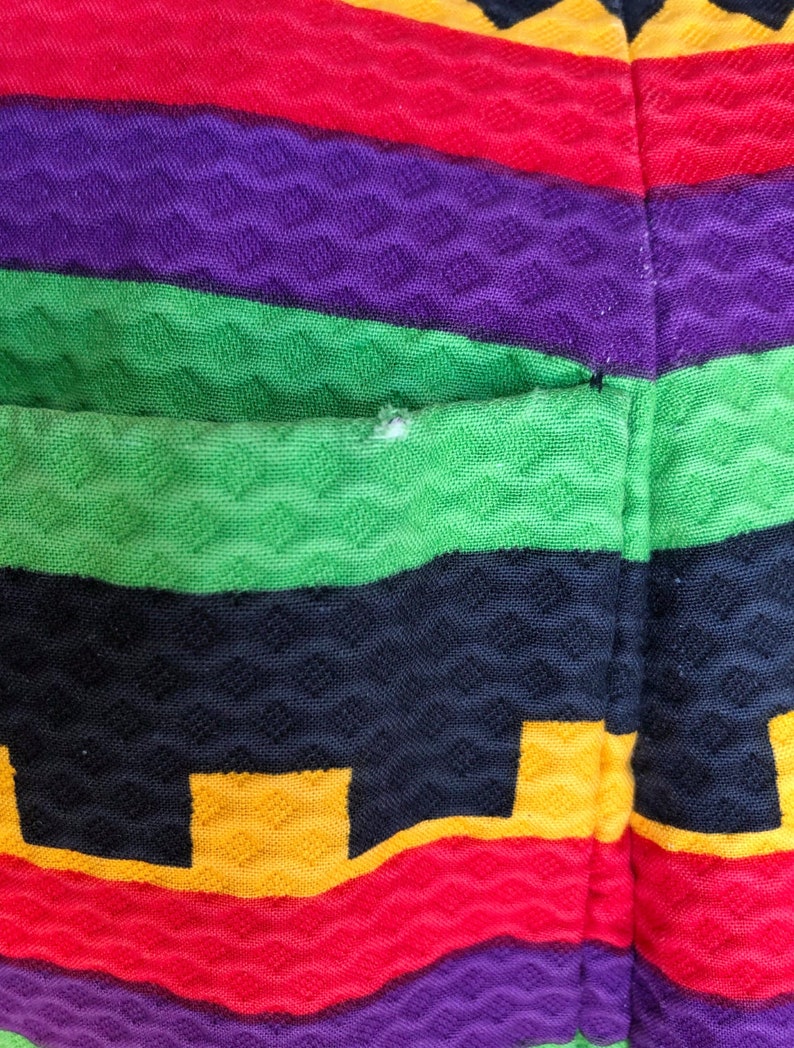 May include: A textured fabric garment featuring horizontal stripes in vibrant shades of yellow, red, purple, green, and black. The material has a raised, diamond-shaped waffle pattern throughout, with a small visible snag on the green stripe.