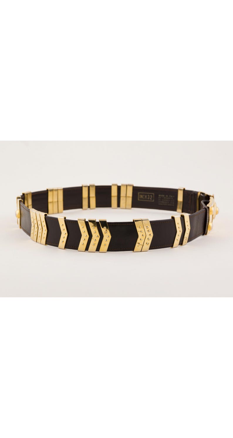 May include: A black leather belt with gold-colored metal accents. The belt features a series of gold rectangular and arrow-shaped embellishments. The belt is approximately 32 inches in length.
