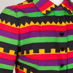 May include: A long-sleeved jacket featuring a textured fabric with horizontal stripes in vibrant purple, bright green, red, and black. The garment includes a classic collar, a front button closure with five black buttons, and a repeating geometric pattern.