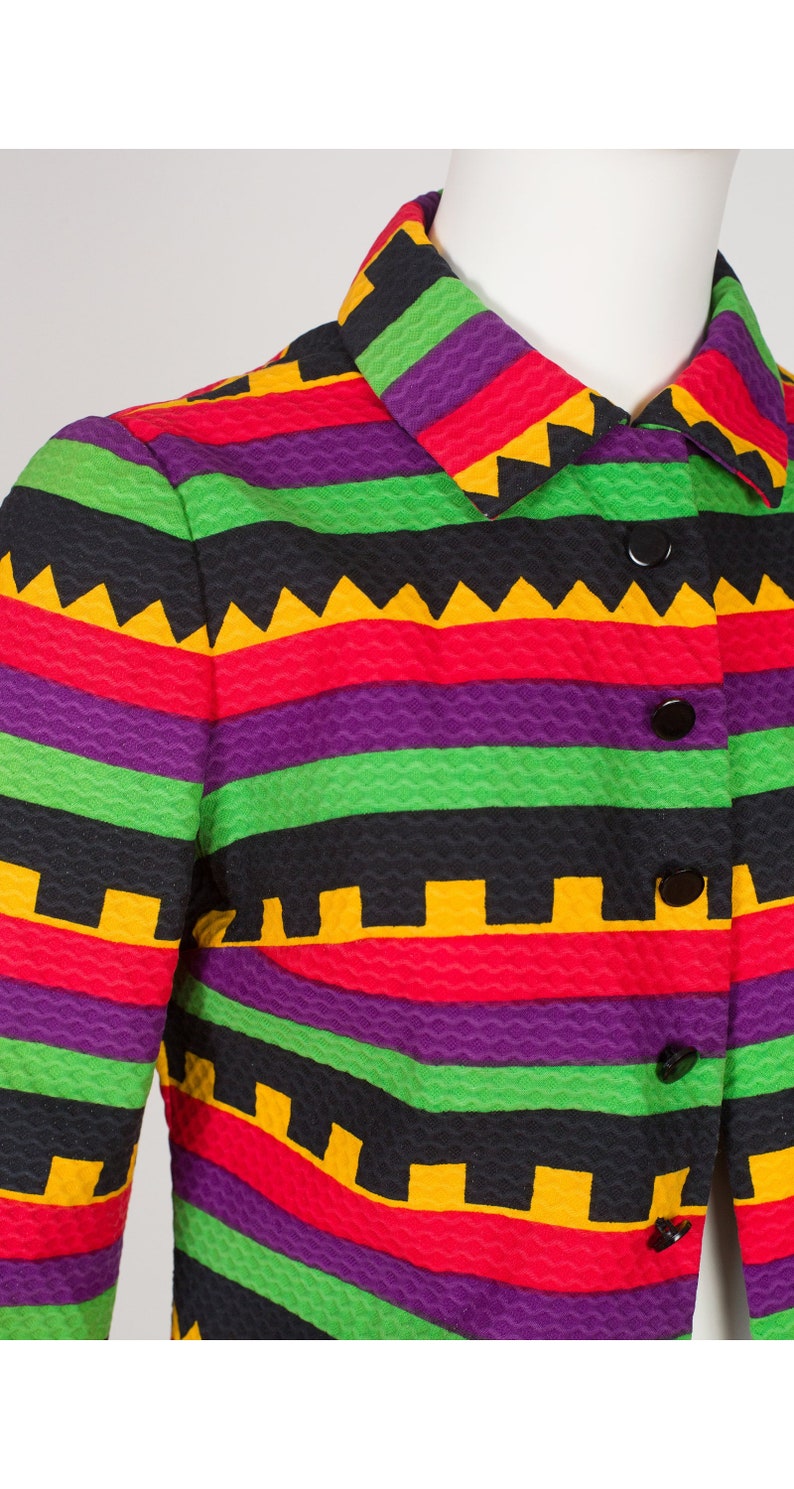 May include: A long-sleeved jacket featuring a textured fabric with horizontal stripes in vibrant purple, bright green, red, and black. The garment includes a classic collar, a front button closure with five black buttons, and a repeating geometric pattern.