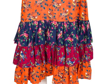 Puede incluir: Una falda escalonada con un estampado vibrante de pájaros y flores. La falda presenta capas de tela naranja, azul marino y burdeos, cada una adornada con pájaros coloridos y delicadas ramas. El diseño de la falda es llamativo y caprichoso.