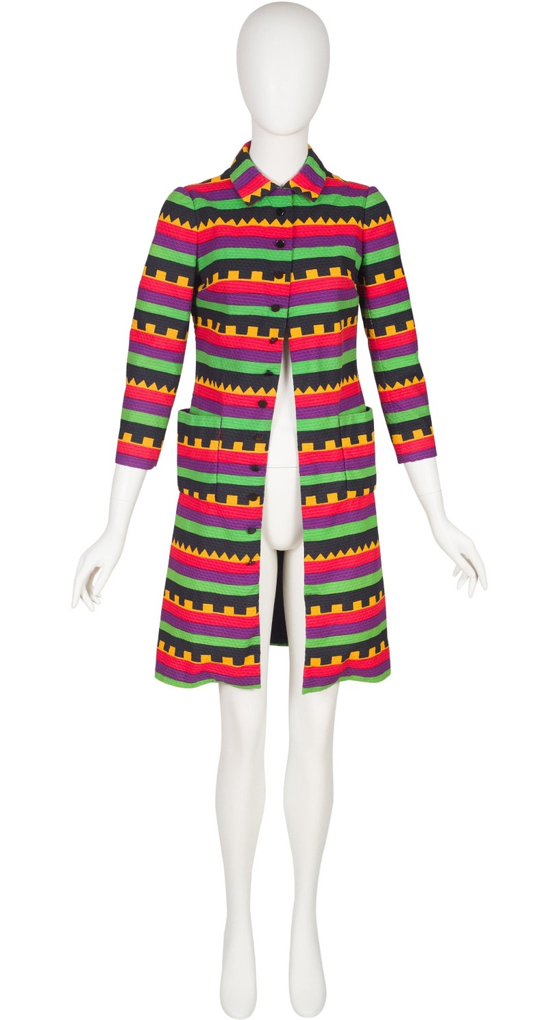 May include: A knee-length coat featuring a vibrant horizontal striped pattern in shades of black, bright green, purple, and yellow. The coat has a classic collar, three-quarter length sleeves, a front button closure, and two rectangular patch pockets.