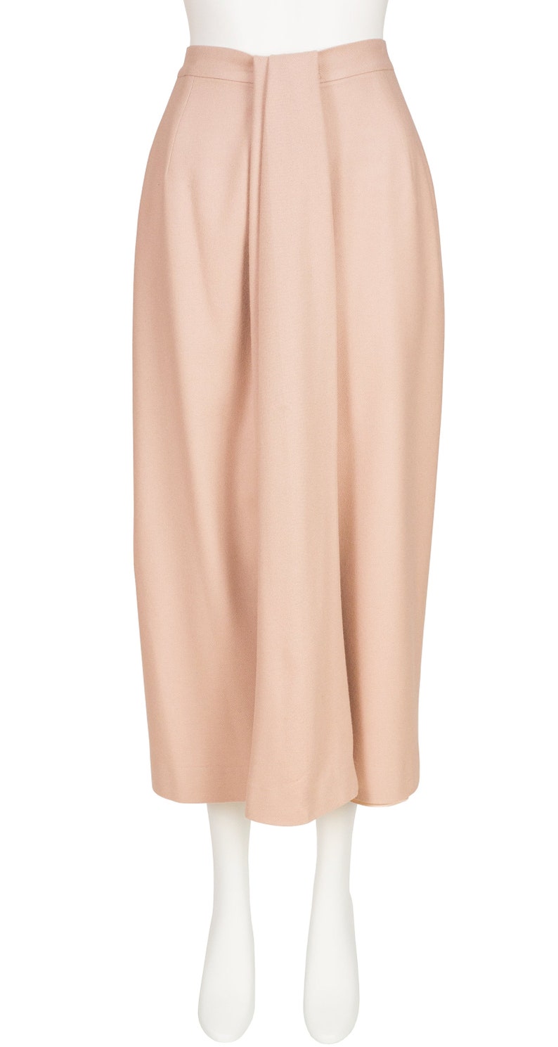 May include: A pale peach skirt with a high waist and a central pleat. The knee-length skirt is made of a smooth, solid fabric. The skirt is displayed on a white mannequin.