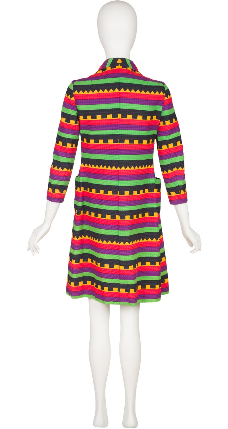 May include: A knee-length coat featuring a vibrant horizontal striped pattern in shades of purple, bright green, red, and yellow. The garment includes a collar, three-quarter length sleeves, and two side pockets, showcasing a textured fabric finish.