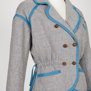 K&ouml;nnte beinhalten: Graue, zweireihige Jacke mit blauen Zierleisten und passenden Kn&ouml;pfen. Die Jacke hat eine geraffte Taille und blaue Zierleisten am Kragen, Revers, Taschen und Manschetten. Die Jacke wird auf einer Schaufensterpuppe pr&auml;sentiert.