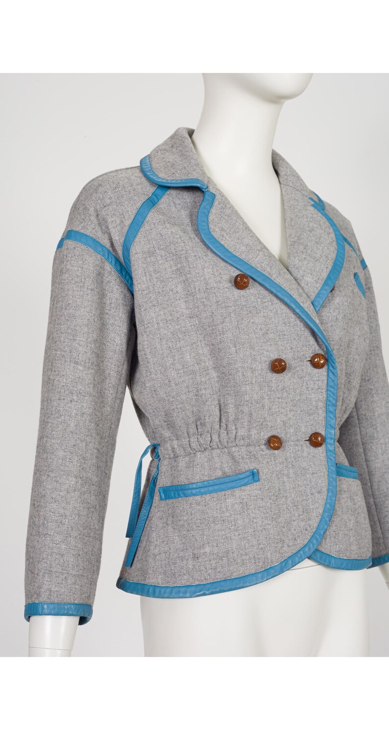 K&ouml;nnte beinhalten: Graue, zweireihige Jacke mit blauen Zierleisten und passenden Kn&ouml;pfen. Die Jacke hat eine geraffte Taille und blaue Zierleisten am Kragen, Revers, Taschen und Manschetten. Die Jacke wird auf einer Schaufensterpuppe pr&auml;sentiert.