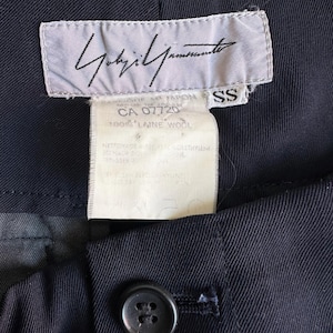 以下が含まれることがあります： 黒いハンドルと「But First, Coffee」のテキストが黒い太字で書かれた白いセラミックマグ。マグは白い表面に置かれています。