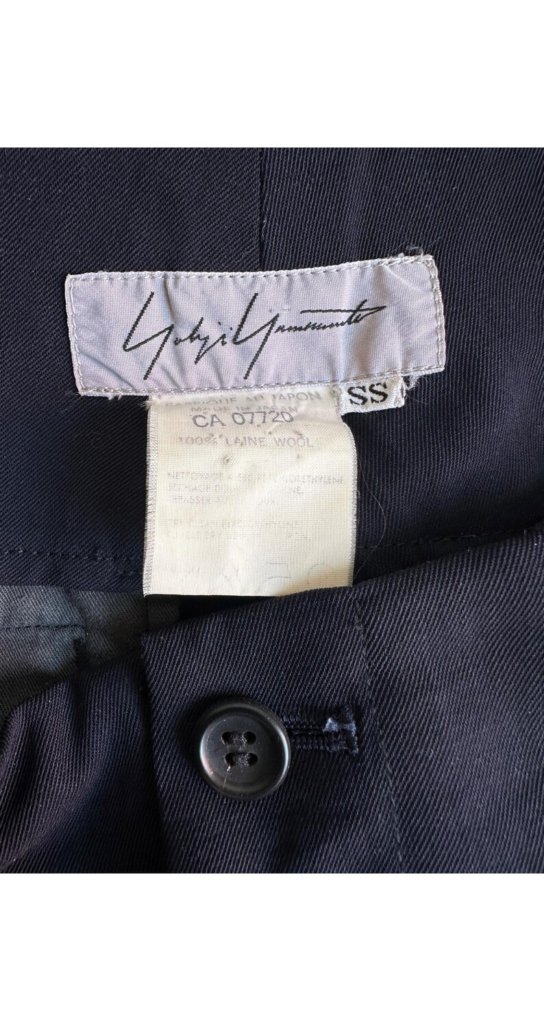 以下が含まれることがあります： 黒いハンドルと「But First, Coffee」のテキストが黒い太字で書かれた白いセラミックマグ。マグは白い表面に置かれています。