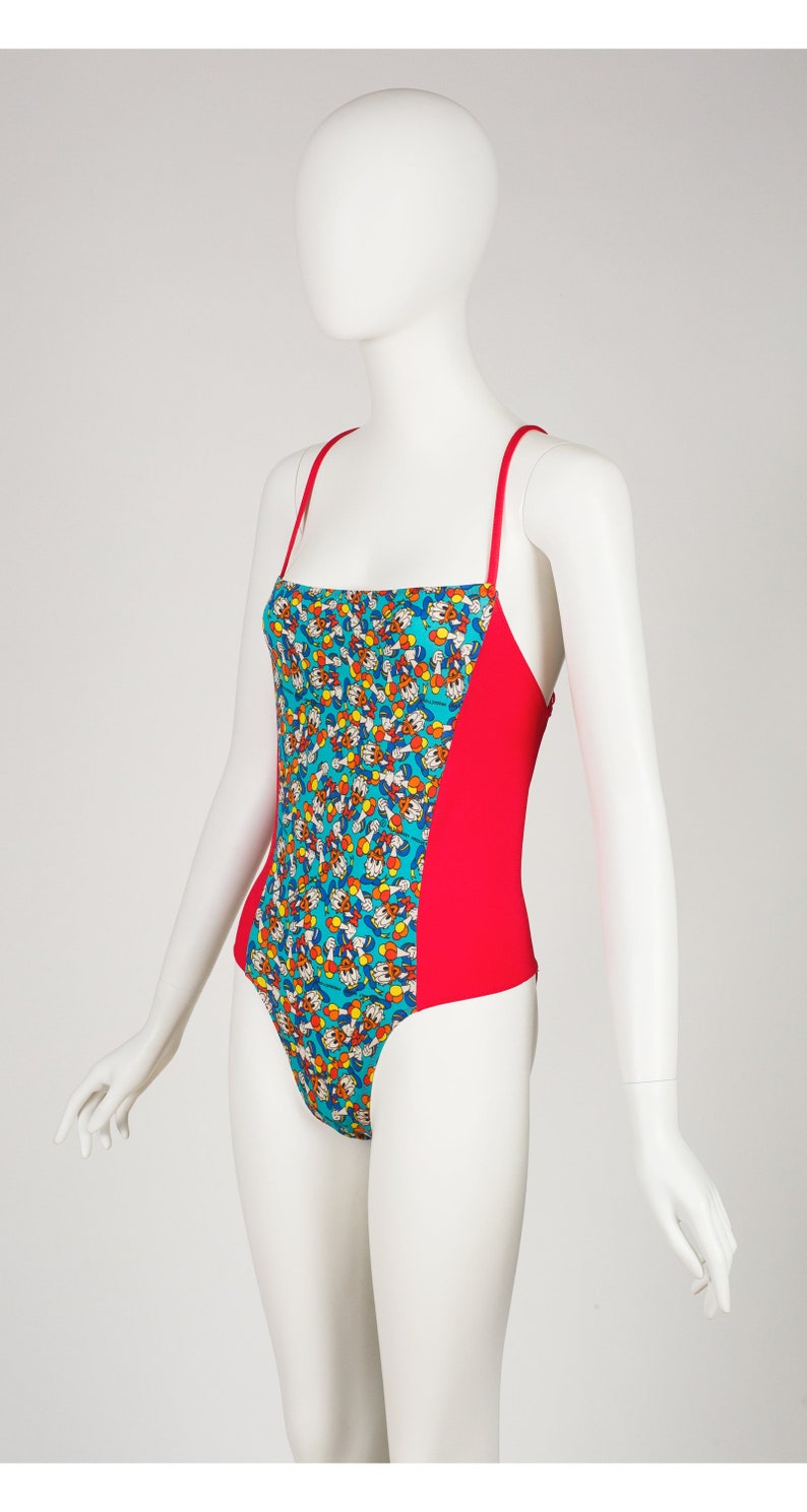 May include: A red and turquoise one-piece swimsuit with a cartoon print of a character with yellow gloves and a red hat.