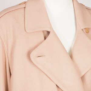 May include: A light beige trench coat is shown in close-up. The coat has a double-breasted design, large buttons, a notched collar, and shoulder epaulets. The fabric looks like a soft, woven material.