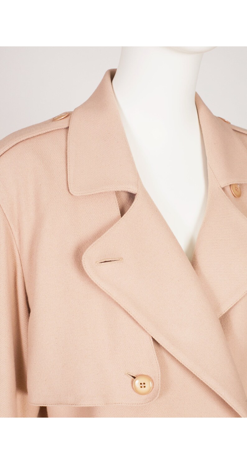 May include: A light beige trench coat is shown in close-up. The coat has a double-breasted design, large buttons, a notched collar, and shoulder epaulets. The fabric looks like a soft, woven material.