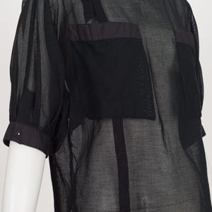 K&ouml;nnte beinhalten: Schwarzes, durchsichtiges Oberteil mit kurzen &Auml;rmeln und rundem Ausschnitt. Das Oberteil hat zwei quadratische Taschen auf der Brust und eine dunkle Paspelierung an Ausschnitt, Taschen und &Auml;rmeln. Die &Auml;rmel haben ein Knopfdetail am B&uuml;ndchen.