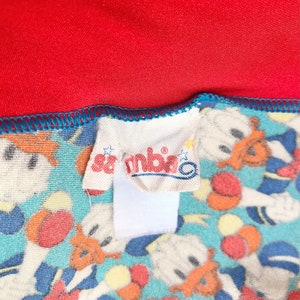 May include: A close-up of a white fabric label with the word "Sambito" printed in red. The label is attached to a blue and white fabric with a pattern of cartoon characters.