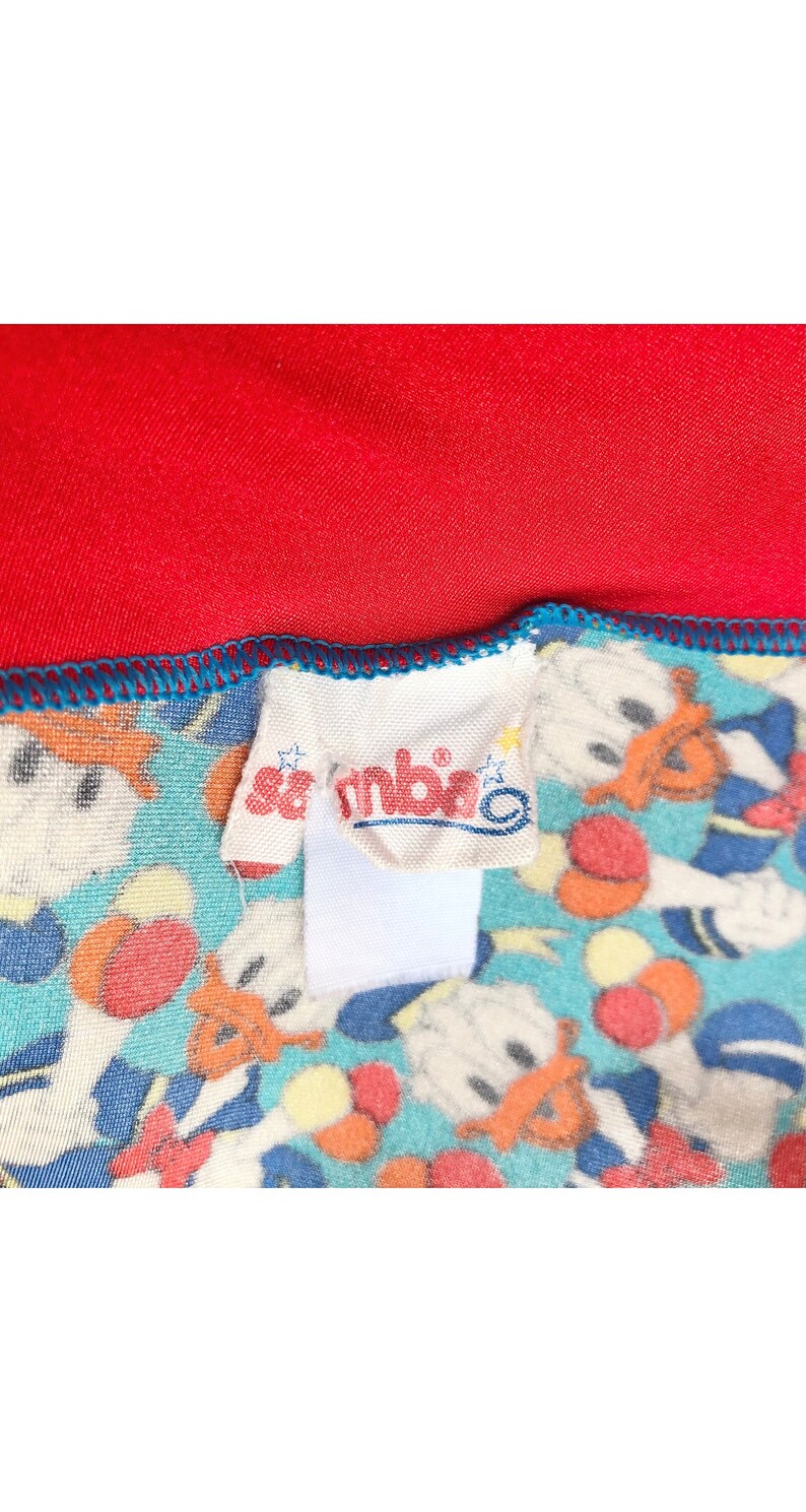May include: A close-up of a white fabric label with the word "Sambito" printed in red. The label is attached to a blue and white fabric with a pattern of cartoon characters.