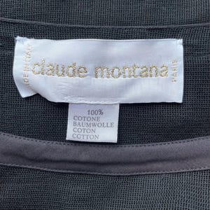 K&ouml;nnte beinhalten: Nahaufnahme eines schwarzen Kleidungsst&uuml;cks mit einem Etikett mit goldfarbenem Schriftzug "claude montana". Das Etikett weist auch "MADE IN ITALY" und "PARIS" auf. Ein kleineres Etikett darunter gibt an, dass das Kleidungsst&uuml;ck aus 100 % Baumwolle besteht.