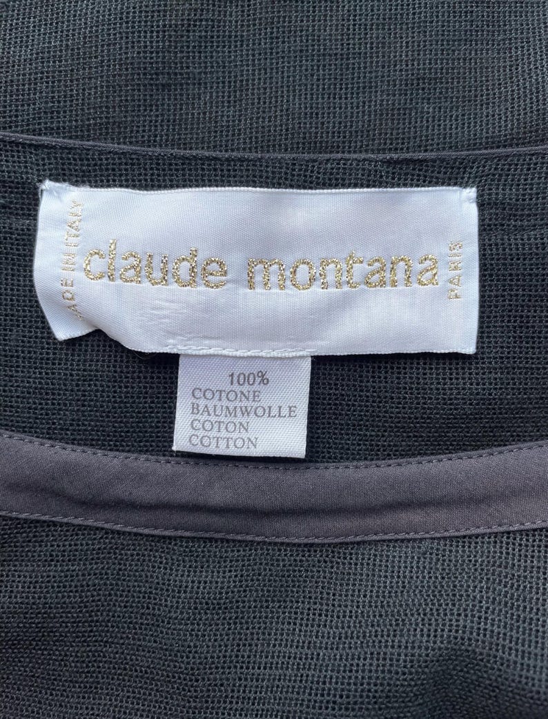 K&ouml;nnte beinhalten: Nahaufnahme eines schwarzen Kleidungsst&uuml;cks mit einem Etikett mit goldfarbenem Schriftzug "claude montana". Das Etikett weist auch "MADE IN ITALY" und "PARIS" auf. Ein kleineres Etikett darunter gibt an, dass das Kleidungsst&uuml;ck aus 100 % Baumwolle besteht.