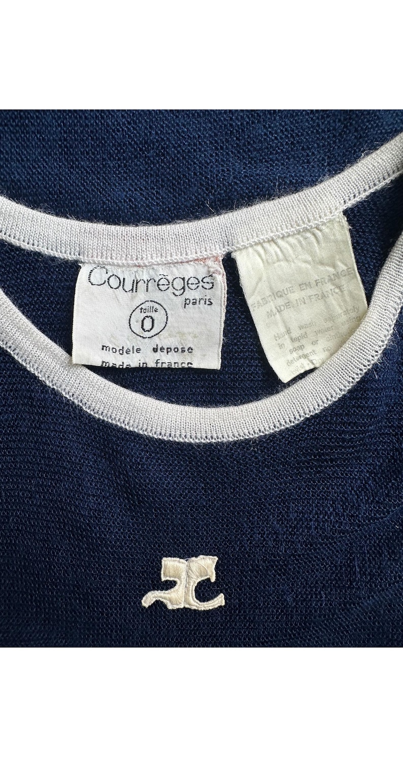 Peut inclure: Haut en maille bleu marine avec une bordure blanche autour de l'encolure et des manches. L'&eacute;tiquette indique "Courr&egrave;ges paris" et "made in france". Un petit logo blanc brod&eacute; est visible en bas.