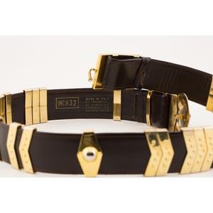 May include: A brown leather belt with gold-colored metal accents. The belt is marked "INCH 32" and has a geometric design. The buckle and other hardware are also gold. The belt is labeled "Made in Italy".