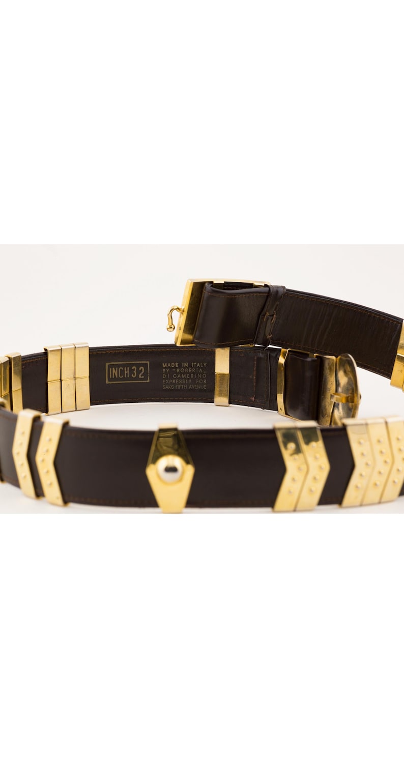 May include: A brown leather belt with gold-colored metal accents. The belt is marked "INCH 32" and has a geometric design. The buckle and other hardware are also gold. The belt is labeled "Made in Italy".