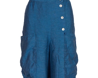 May include: Teal-colored linen pants with large front pockets and a button closure. The pants have a high waist and a relaxed fit. The buttons are silver. The fabric has a textured appearance.
