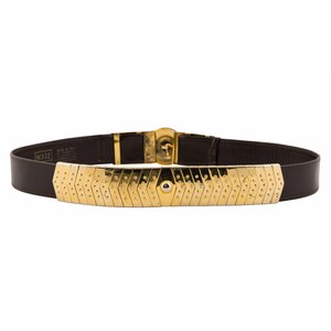 May include: A brown leather belt with a gold-colored metal front piece. The metal piece has a geometric design with small, evenly spaced holes. The belt buckle and hardware are also gold-colored. The belt is labeled "NC#32".