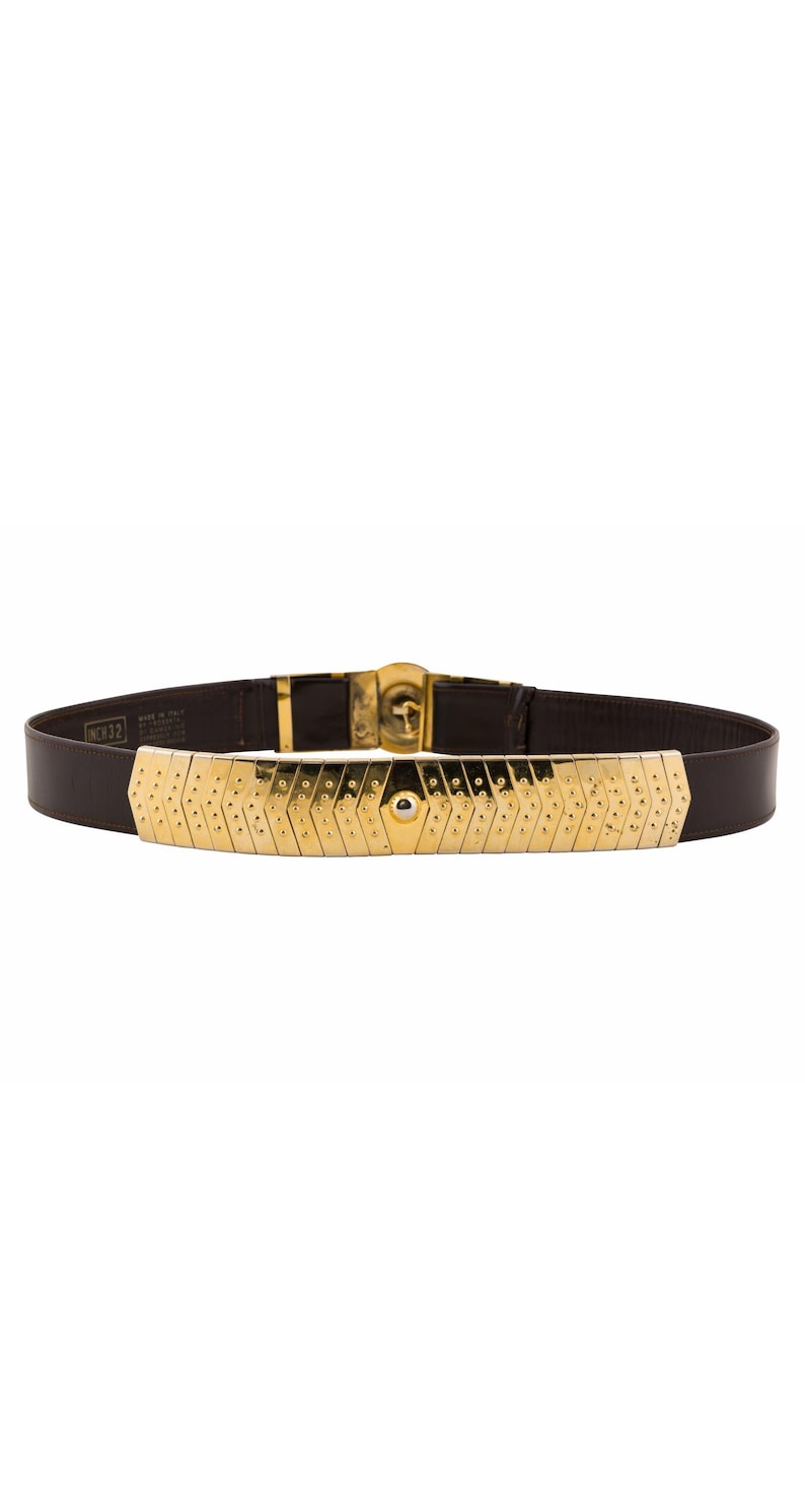 May include: A brown leather belt with a gold-colored metal front piece. The metal piece has a geometric design with small, evenly spaced holes. The belt buckle and hardware are also gold-colored. The belt is labeled "NC#32".