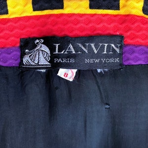 May include: A black garment label with white text reading LANVIN PARIS NEW YORK and a graphic of a person. Below the label is a small white tag with the red number 8. The fabric above the label features horizontal stripes of yellow, black, red, and purple.